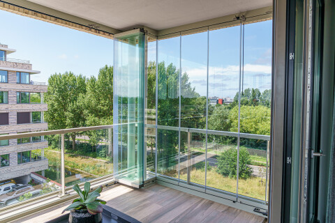 Balkon in Katwijk met glazen vouwbeglazing deels open in de hoek, met uitzicht op bomen en omliggende appartementen