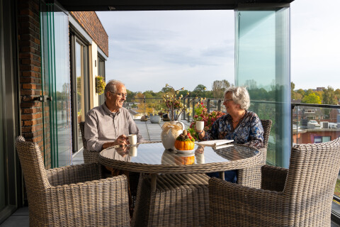 Twee ouderen zitten aan een ronde tuintafel op een balkon met glazen schuifwanden, met koffie en uitzicht over de omgeving